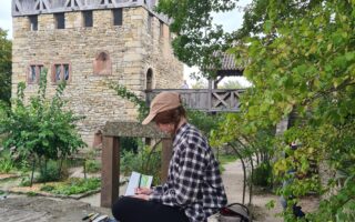 Eine Frau sitzt malend auf einer Mauer und schaut draußen auf ein Haus